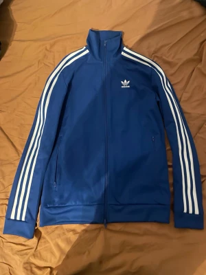 Blå Adidas track jacka  - Säljer en klassisk blå Adidas track jacket med vita ränder längs ärmarna och dragkedja framtill. Jackan har hög krage, två fickor med dragkedja och Adidas-logga på bröstet. Perfekt för en sportig och avslappnad stil.