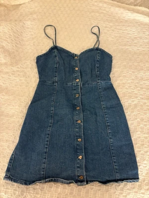 Kort jeansklänning med knappar - Säljer en snygg kort jeansklänning med tunna axelband och knappar framtill. Klänningen har en figurnära passform och är tillverkad i klassiskt blått denimtyg. Sparsamt använd😍storlek M men sitter som en S