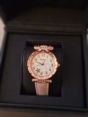 Beige klocka med roséguld och blommor - Snygg klocka från Inschiv med beige band och roséguldfärgat ur. Urtavlan har blommigt mönster och glittriga detaljer runt kanten. Bandet är i syntetmaterial med vit söm och klassiskt spänne. Perfekt accessoar för att lyfta din stil.