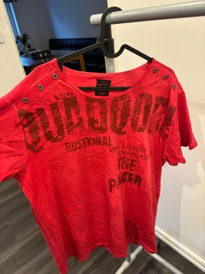 Röd Rusty Neal t-shirt med tryck XL - Cool röd t-shirt från Rusty Neal i storlek XL med stora tryck både fram och bak. T-shirten har korta ärmar, rund halsringning och snygga knappdetaljer på axlarna. Materialet är mjukt och skönt, perfekt för en avslappnad streetstil.