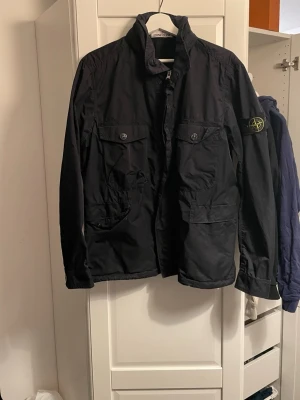 Stone island overshirt  - Säljer nu min stone island overshirt jacka då den blivit för liten, jackan är marinblå i storlek M, jackan har ett par år på nacken och använts flitigt så visst slitage finns men inga hål eller dylikt. Finns även luva som går att rulla ut i nacken