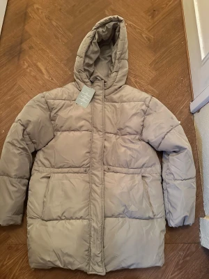 Beige pufferjacka med huva - Snygg beige pufferjacka från H&M med huva och dragkedja framtill. Jackan har två stora fickor och quiltad design som ger en cool och avslappnad look. Perfekt för kalla dagar när du vill hålla dig varm och stilren.