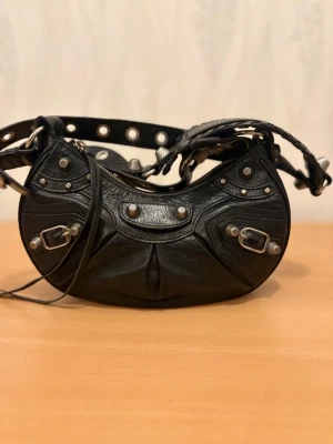 Balenciaga svart axelväska i skinn - Cool svart axelväska från Balenciaga i äkta skinn med silvriga nitar och spännen. Väskan har en unik halvmåneform, grov dragkedja och detaljerad design med flera metalldetaljer. Perfekt för dig som vill sticka ut med en ikonisk designerbag.