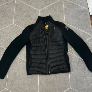 Svart pufferjacka från Parajumpers S - Svart pufferjacka från Parajumpers i storlek S. Jackan har quiltad front, långärmade släta ärmar och en gul detalj vid kragen. Dragkedja framtill och diskret logga på ärmen. Perfekt för dig som vill ha en clean och sportig look.