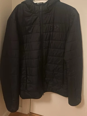 Svart pufferjacka EA7 Emporio Armani - Svart pufferjacka från EA7 Emporio Armani med huva och snyggt, glansigt logotryck på bröstet. Jackan har quiltad design, dragkedja framtill och två sidofickor med dragkedja. Perfekt för dig som vill ha en stilren och modern look.