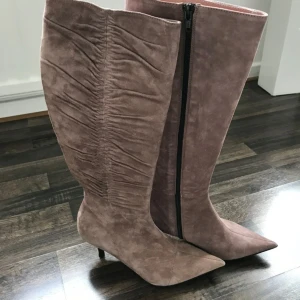 Beige rosa mockastövlar med spetsig tå - Säljer ett par höga stövlar i beige rosa mocka med snygg rynkad detalj längs skaftet och spetsig tå. Stövlarna har smal klack och dragkedja på insidan för enkel påtagning. Perfekt för dig som gillar stilrena och trendiga boots med lite edge.