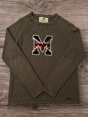 Brun stickad tröja Morris med M-tryck - Snygg brun stickad tröja från Morris med ett stort M på bröstet, dekorerat med Union Jack-mönster i rött, blått och vitt. Tröjan har långärmad passform och rund hals. Diskret Morris-brodyr vid nederkanten. Perfekt för dig som gillar klassisk stil med en twist.