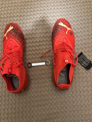 Röda fotbollsskor från Puma - Säljer ett par grymt snygga fotbollsskor från Puma i röd färg med guldiga detaljer och logga. Skorna har snörning, spetsig tå och metall-dobbar för extra grepp på planen. Ovandelen är i syntetmaterial med ventilerande mesh och coolt prickigt mönster.
