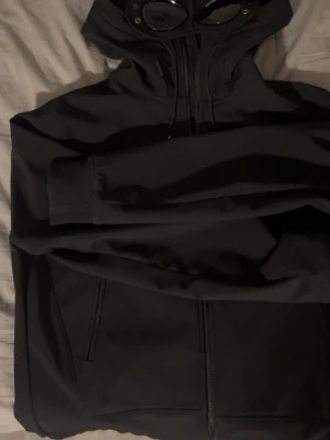 Svart vindjacka från C.P. Company - Svart vindjacka från C.P. Company med huva och ikoniska goggles-detaljer. Jackan har dragkedja framtill, två sidofickor med dragkedja och justerbar huva med snörning. Materialet är syntet och designen är clean och modern, perfekt för dig som gillar streetwear.