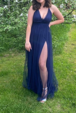 Marinblå balklänning  - Säljer nu min fina balklänning som jag hade i nian. Den är marinblå med v-ringning och tunna axelband som korsas i ryggen. Klänningar har en slits på varje sida och en kjol i tyll över! Perfekt för student eller bal!