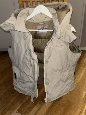 Beige dunväst från Henri Lloyd - Snygg beige dunväst från Henri Lloyd med huva och dragkedja samt knappar framtill. Västen har broderad logga på bröstet och en cool detalj med brittisk flagga på sidan. Fodret är i en matchande beige ton och materialet känns slitstarkt och mjukt.