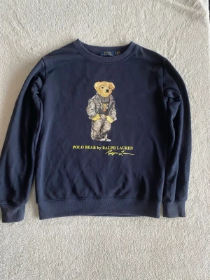 Mörkblå Polo Bear tröja Ralph Lauren - Säljer en mörkblå sweatshirt från Ralph Lauren i storlek S med ikoniskt Polo Bear-tryck på bröstet. Tröjan har rund hals, ribbade muddar och är tillverkad i mjuk bomull. Perfekt för dig som gillar streetstyle och klassiska märken.