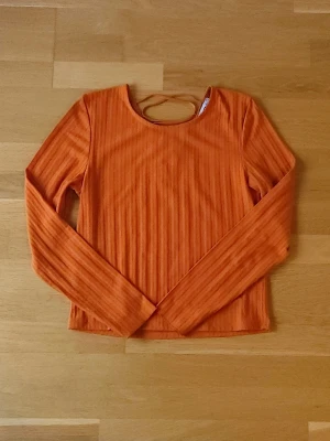 Orange topp - Snygg ribbad orange topp med långa ärmar och snörningsdetalj i ryggen. Toppen har en rund halsringning fram och ett v-format snörat parti bak som ger en snygg look.