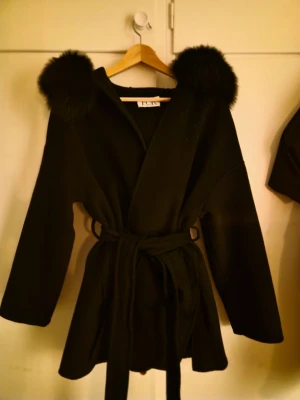 Svart pälsjacka från FUR Stockholm - Snygg svart pälsjacka från FUR Stockholm med äkta päls runt huvan.Perfekt för dig som vill ha en stilren och varm look under höst och vinter. Jackan har stora fickor framtill. Kappan består av 90% ULL och 10% kashmir, pälskragen består av 100% rävpäls. Köpte den orginalt för 3000kr och har knappt kommit till används