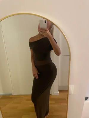 Brun lång klänning  - Säljer en snygg brun långklänning med offshoulder-desig vid ena axeln  och smal passform. Klänningen har ett transparent meshparti i midjan som ger en cool detalj. Materialet är mjukt och stretchigt, vilket gör den riktigt bekväm och följsam. Perfekt för dig som gillar stilrena och trendiga plagg. Helt ny pris lapp kvar.
