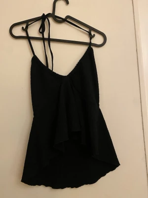 Svart linnetopp med volang - Säljer en svart linnetopp med smala axelband som knyts i nacken. Toppen har en snygg volang framtill och är längre baktill för en cool asymmetrisk look. Perfekt för dig som gillar stilrena och trendiga plagg med lite edge.