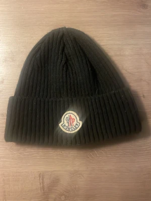 Svart ribbad mössa från Moncler - Svart ribbstickad mössa från Moncler med klassisk uppvikt kant och broderad logotyp framtill. Snygg och enkel design som passar till de flesta jackor. Materialet är mjukt och värmande, perfekt för kyliga dagar.