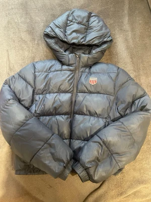 Mörkblå pufferjacka från GANT🫶🏻 - Säljer en mörkblå pufferjacka från GANT med quiltad design och huva. Jackan har dragkedja framtill, två fickor och GANT-logga på bröstet. Perfekt för kalla dagar och riktigt snygg till streetwear-stilen. Den har blivit lagad (se bild fyra) och därav priset! Men jättesnygg!🤍