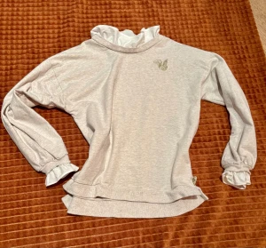 Beige långärmad tröja från POMPdeLUX - Säljer en beige långärmad tröja från POMPdeLUX med volangkrage och volangdetaljer vid ärmsluten. Tröjan har ett broderat motiv på bröstet och en liten hjärtformad etikett vid nederkanten. Mjuk bomullskvalitet och loose passform. 95% ekologisk bomull 5% elastan