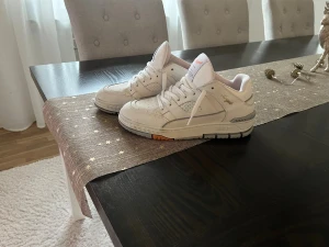 Vit sneaker Axel Arigato - Säljer ett par cleana vita sneakers från Axel Arigato med gulddetaljer och klassisk siluett. Skorna har perforerad tå, snörning och robust sula. Tillverkade i skinn för en lyxig känsla och har diskret logga på sidan.