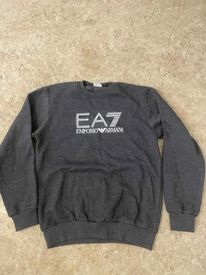 Mörkgrå EA7 Emporio Armani sweatshirt - Snygg mörkgrå sweatshirt från EA7 Emporio Armani med stort vitt logotryck på bröstet. Klassisk rund halsringning, ribbade muddar och midja. Perfekt för dig som gillar stilrena och sportiga vibes.