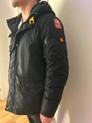 Svart Parajumpers parkajacka herr - Svart parkajacka från Parajumpers med huva, flera fickor och gul detalj vid halsen. Jackan har patch på ärmen och dragkedja framtill. Insidan är fodrad och materialet är slitstark polyester. Har även päls till luvan som ingår!
