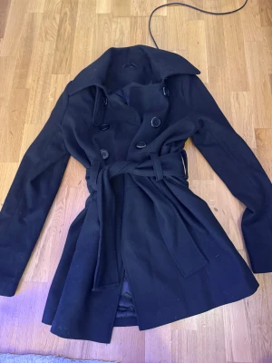 Svart trenchcoat med bälte - Snygg svart trenchcoat med klassisk krage och dubbelknäppning framtill. Jackan har ett brett bälte i midjan som knyts och stora svarta knappar. Långärmad och med en stilren, tidlös look som passar perfekt till höst och vinter.
