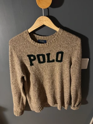 Beige stickad tröja Polo Ralph Lauren - Stickad beige tröja från Polo Ralph Lauren i storlek XL men i barnstorlek. Tröjan har grönt POLO-tryck framtill,  rund hals och ribbade muddar vid ärmslut och nederkant. Det är äkta ralph lauren köpt i butik. Perfekt för dig som gillar klassisk stil med en sportig touch. Obs kraken är lite utdragen men inget jätte stort.