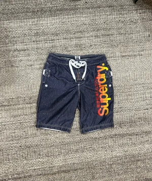 Superdry blå shorts med snörning - Snygga mörkblå shorts från Superdry med vit snörning i midjan och stort Superdry-tryck i orange och gult längs benet. Shortsens material är polyester och de har vita kontrastsömmar samt praktiska fickor på sidorna.