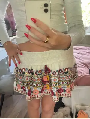 Superfin kortkjol med broderade blommor i flera färger och volangkant. Kjolen är vit i grunden och har detaljer i rött, rosa, gult, grönt och blått. Den har snörning med tofsar framtill och är perfekt för sommarens alla upplevelser. Söt och färgglad. Skit snygg unik kjol. Nervikt på bilden. Be gärna om mer bilder vid intresse.