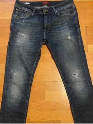 Mörkblå jeans från Jack & Jones - Snygga mörkblå jeans från Jack & Jones med slitna detaljer och klassisk femficksdesign. Jeansen har en smal passform och är tillverkade i stretchigt denim för extra komfort. Perfekta för dig som gillar en modern och avslappnad stil.
