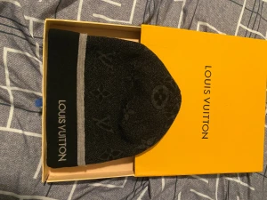 Svart Louis Vuitton mössa med logomönster - Exklusiv svart mössa från Louis Vuitton med diskret logomönster och grå rand. Mössan är stickad i mjukt material och har broderad Louis Vuitton-text på kanten. Perfekt accessoar för dig som vill ha en lyxig och stilren look.