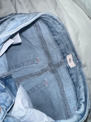 Ljusblå jeans med hål från Nelly - Säljer ett par ljusblå jeans från Nelly med breda ben och stora hål vid knäna. Jeansen har klassisk femficksdesign, röd broderad detalj på bakfickorna och slitna kanter. Perfekt för dig som gillar en chill och edgy stil.