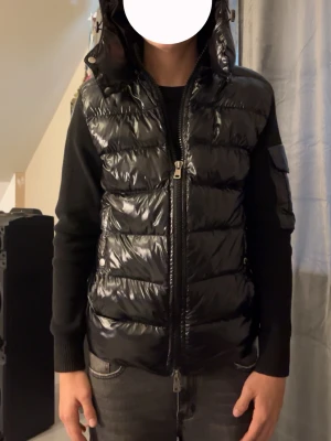 Svart dunväst från Moncler - Säljer en svart, glansig dunväst från Moncler med huva och dragkedja framtill. Västen har quiltad design och coola detaljer som fickor med tryckknappar. Perfekt för att addera en lyxig touch till din outfit. Moncler-logga och etikett med serierutor på insidan.