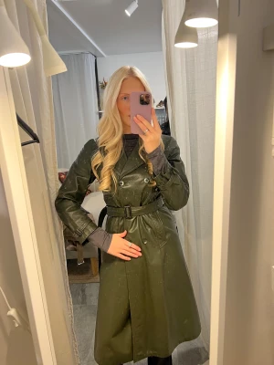 Mörkgrön trenchcoat i skinn - Säljer en riktigt snygg mörkgrön trenchcoat i skinn, med bälte i midjan och dubbla knapprader. Är en storlek M och jag kan ha denna 