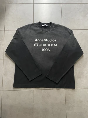 Svart långärmad tröja Acne Studios - Svart långärmad tröja från Acne Studios med vit text 'Acne Studios STOCKHOLM 1996' på bröstet. Tröjan har rund halsringning och en avslappnad passform. Tillverkad i mjuk bomull, perfekt för en stilren och enkel look.