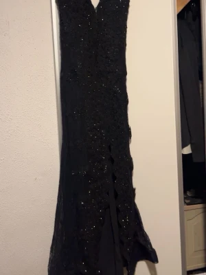 Svart långklänning med spets och glitter - Elegant svart långklänning med v-ringning och spetsdetaljer över hela klänningen. Klänningen har glittriga paljetter som ger extra lyxig känsla och en slits framtill. Dragkedja i ryggen för enkel påklädning. Perfekt för bal eller festliga tillfällen. Använt bara en gång köpte den kostade 4500kr.