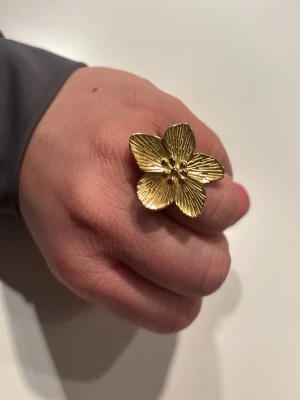 Guldfärgad blomring med struktur - Snygg ring i guldfärg med en stor blomma som har fem blad och detaljerad struktur. Ringen är justerbar och har en cool, glansig finish som verkligen sticker ut. Perfekt för dig som gillar statement-smycken och vill ha något unikt.