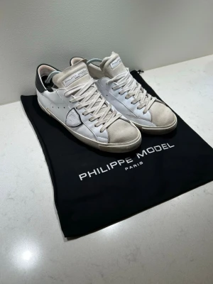 Philippe Model PRSX sneakers - Snygga vita Philippe Model sneakers med beige mockadetaljer på tån och plös, samt svart hälparti. Skorna har klassisk snörning och rund tå. Tillverkade i skinn och mocka för en lyxig känsla. Perfekta för dig som gillar stilrena och trendiga sneakers. Det ingår dustbag med skorna. Dem är sparsamt använda. Det är bara att höra av sig vid frågor eller funderingar!