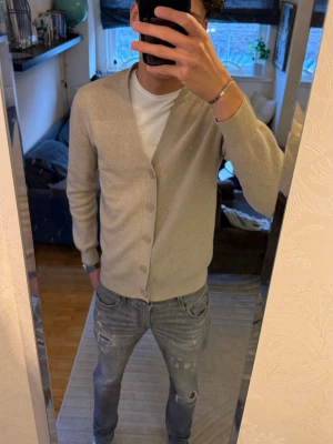 Beige kofta - Snygg beige kofta från Primark i storlek medium. Koftan har en klassisk v-ringad design med fem knappar framtill och långa ärmar. Materialet är mjukt och stickat, perfekt för lager-på-lager. Enkel och stilren look som funkar till jeans och t-shirt.