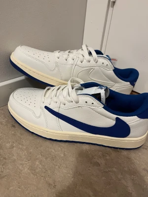 Nike Air Jordan 1 Low Travis Scott Vit/Blå - Nike Air Jordan 1 Low Travis Scott i vitt och blått med omvänd blå Swoosh, broderad Air Jordan Wings-logga och Cactus Jack-detalj på hälen. Skorna har perforerad tå, platt sula och extra skosnören i rosa och blå. Tillverkade i skinn med blå textilfoder.