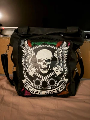 Svart axelväska cafe racer  - Cool svart axelväska i skinnmittation med stort tryck av dödskalle, vingar och texten 'Cafe Racer' på framsidan. Väskan har både handtag och justerbar axelrem, samt dragkedjeficka på baksidan och många fack inuti. Färgdetaljer i rött, grönt och lila på framsidan.
