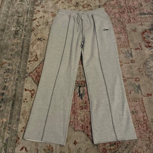 Grå mjukisbyxor från Corteiz - Snygga ljusgrå mjukisbyxor från Corteiz med svarta kontrastsömmar och broderad logga på framsidan. Byxorna har elastisk midja med snörning och raka, vida ben. Tillverkade i mjuk bomull för maximal komfort. Bakficka med patchdetalj.