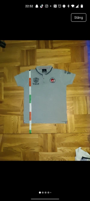 Grå polo t-shirt Ebound Denim M - Snygg grå polo t-shirt från Ebound Denim med broderade och tryckta New York/Manhattan detaljer fram och bak. Kragen har blå och vita ränder, och tröjan har korta ärmar samt knappar framtill. Materialet är mjuk bomull, perfekt för en chill streetstyle.