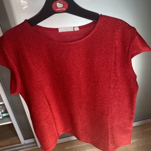Röd glittrig topp från KappAhl - Snygg röd topp från KappAhl med glittrigt tyg. Modellen är kortärmad och har en rund halsringning. Perfekt till nu till julen.