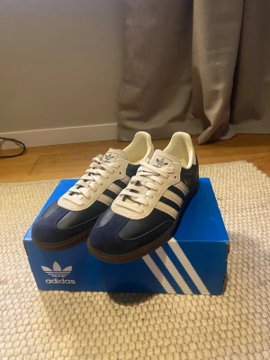 adidas Samba OG Aurora Ink Cream White - adidas Samba OG Aurora white. Ovandel i skinn och mocka, vit plös och snörning. Brun gummisula med Adidas-logga undertill. Helt ny och oandvänd. äktehetsverifierad av hypeboost. Säljs då de inte passar.