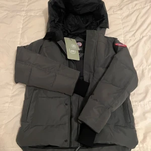 Mörkgrå dunjacka från Canada Goose - Snygg mörkgrå dunjacka från Canada Goose med huva och klassisk logga på ärmen. Jackan har quiltad design, dragkedja framtill och ribbade muddar vid ärmslut. Perfekt för kalla dagar och har en clean, urban look.