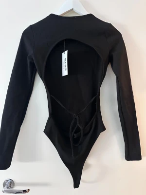 Svart långärmad bodysuit från NA-KD - Svart bodysuit från NA-KD med lång ärm och rund hals. Baksidan har en stor öppen rygg och snygg snörning som knyts i midjan. Modellen är tight och tillverkad i mjuk bomull med lite stretch för en skön passform. Perfekt för att skapa en cool och trendig look.