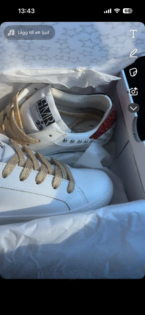 SMR23 vita sneakers med röda glitterdetaljer - Snygga vita sneakers från SMR23 med röda glitterdetaljer och silvriga nitar på sidan och hälen. Skorna har guldig snörning och en cool blixt i rött glitter på sidan. Köpte dom i Norge i september och de är knappt använda. Köpta för 2200 norska kronor😊
