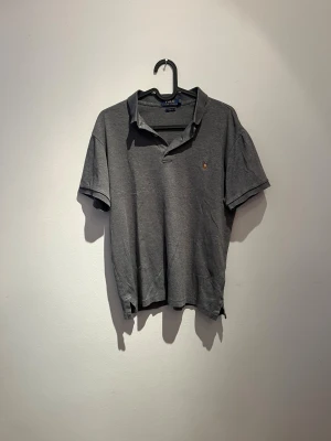 Grå pikétröja Polo Ralph Lauren M - Grå pikétröja från Polo Ralph Lauren i slim fit med klassisk krage och korta ärmar. Tröjan har en liten broderad logga på bröstet och knappar vid halsen. Tillverkad i mjuk bomull, perfekt för en clean och stilren look. Tyvärr ett litet hål vid knapparna men inget som märks